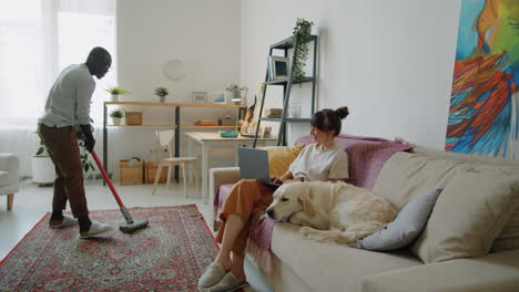 Black-Man-Vacuuming-Floor-while-Wife-and-Dog-Resting-on-Sofa